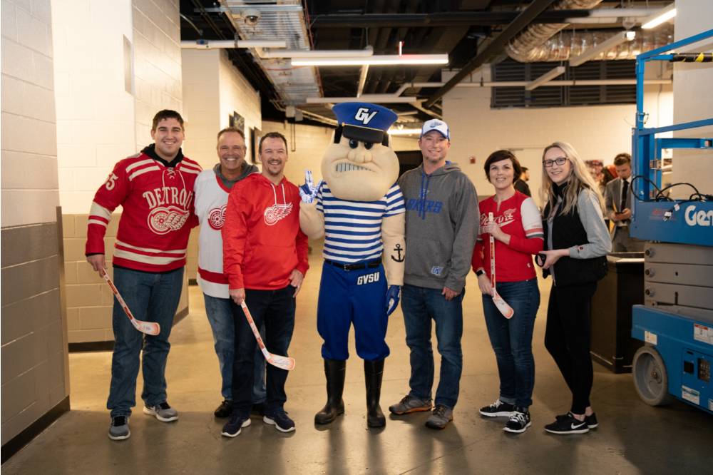 Six alumni smile for a photo together with Louie the Laker at the Detroit Red Wings GVSU Night